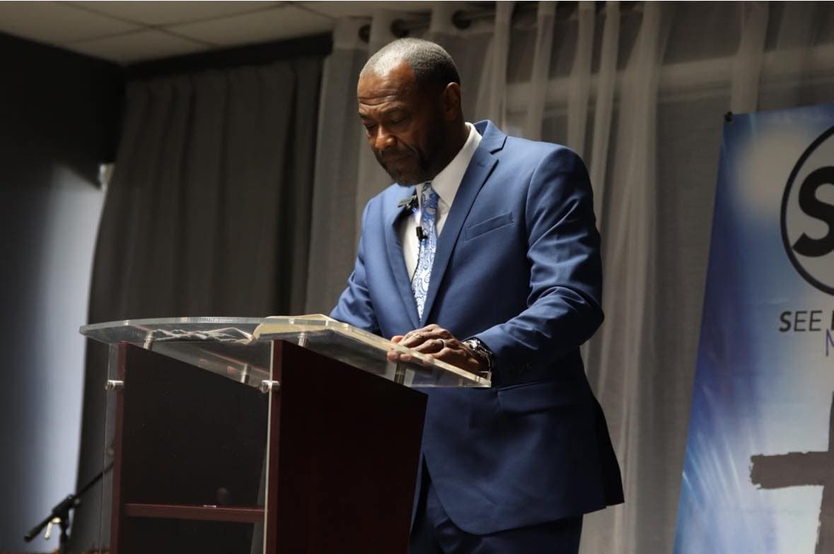 A pastor preaching from the pulpit at See His Face Ministries with his hand resting on an open Bible